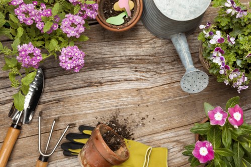 The image displays a rustic wooden outdoor table with various gardening tools and potted plants arranged on its surface. In the top right corner, there is a gray metal watering can with a long spout, alongside a small terracotta pot containing soil and a few decorative wooden plant labels. To the left, a pair of pruning shears with black handles and a three-pronged hand rake are positioned on the table. Bright pink and purple flowering plants, possibly petunias or similar garden flowers, are in pots and placed around the tools, adding vibrant colour and natural contrast to the scene. A small, empty terracotta pot with soil spilling from it sits near the centre, with a pair of yellow gardening gloves partially underneath it. The setup suggests outdoor gardening activities, with fresh flowers and essential tools indicating ongoing or upcoming garden maintenance work typical of professional lawn mowing and landscaping services in Brompton or nearby areas. The weather appears clear, with bright lighting highlighting the fresh soil and blooming flowers, creating an inviting and well-maintained garden ambiance suitable for professional garden care descriptions.