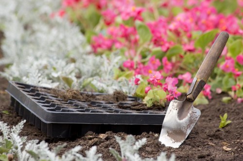 A close-up view of a gardening scene in a front or backyard garden, featuring a black plastic seed tray resting on rich, dark soil, with some dirt and plant debris on top. A small garden trowel with a wooden handle and a metal blade is partially embedded in the soil, suggesting ongoing planting or soil aeration work. Surrounding the tray and trowel, there are various vibrant flowering plants, including clusters of pink and white flowers, possibly begonias or similar seasonal blooms, providing a colourful background. The area appears well-maintained and landscaped, with a focus on preparing the soil for planting. The lighting indicates a clear, bright day, suitable for outdoor gardening activities in Brompton or nearby areas. This scene reflects professional gardening and lawn care practices, consistent with the services offered by Lawn Mowing Brompton, highlighting the preparation and planting stages typical in garden maintenance and landscaping projects.