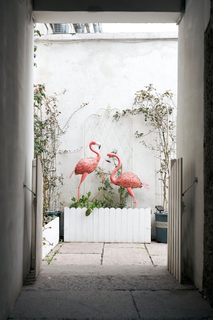 A courtyard garden area surrounded by multi-story apartment buildings with white facades, accented by orange balcony trims and black wrought iron railings. The garden features lush greenery, including